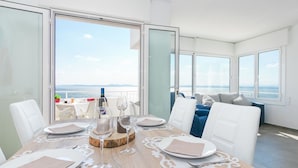 A dining area with a table set for four, large windows offering a view of the sea, and a balcony with seating.