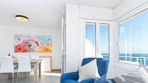 A modern living room with a large window, a dining area with a table and chairs, and a colorful abstract painting on the wall.