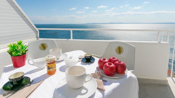A table set with white tableware, a red potted plant, and a bottle of honey on a balcony with a sea view.