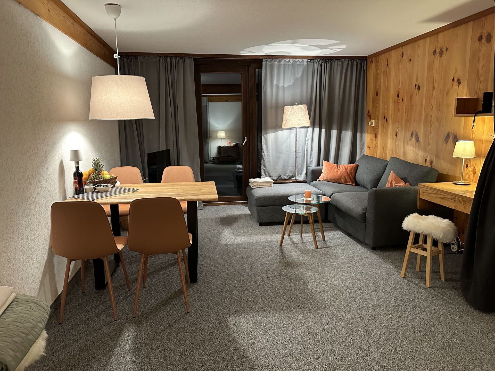 A modern living room with a wooden accent wall, a grey sofa, a dining table with chairs, and a small round coffee table.