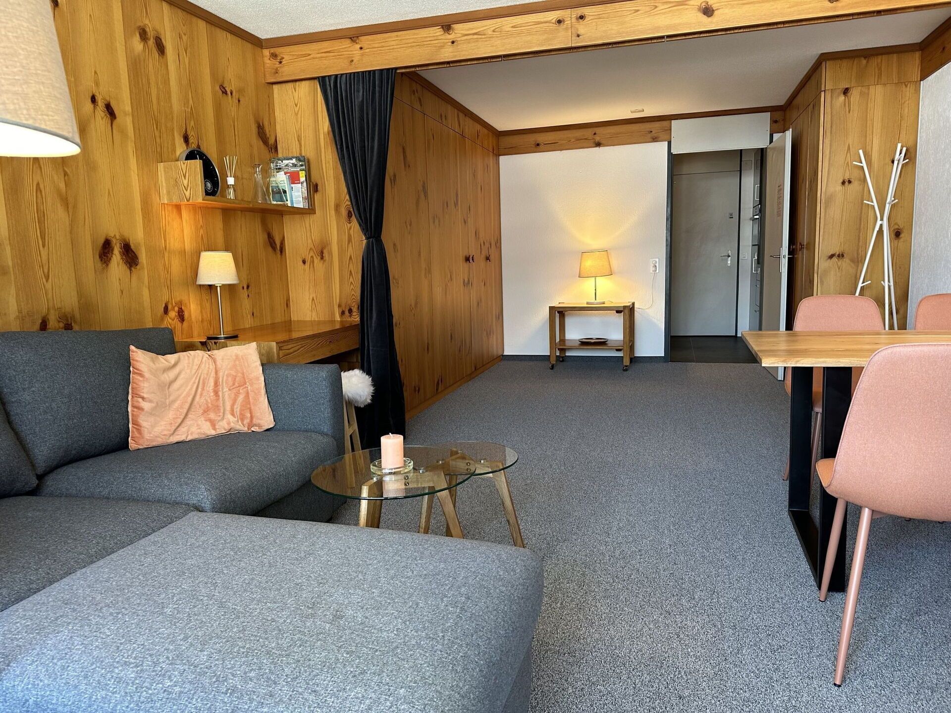 Ett mysigt vardagsrum med en grå soffa, ett litet glasbord med ett ljus, ett träbord med en lampa och en matplats med ett träbord och stolar.