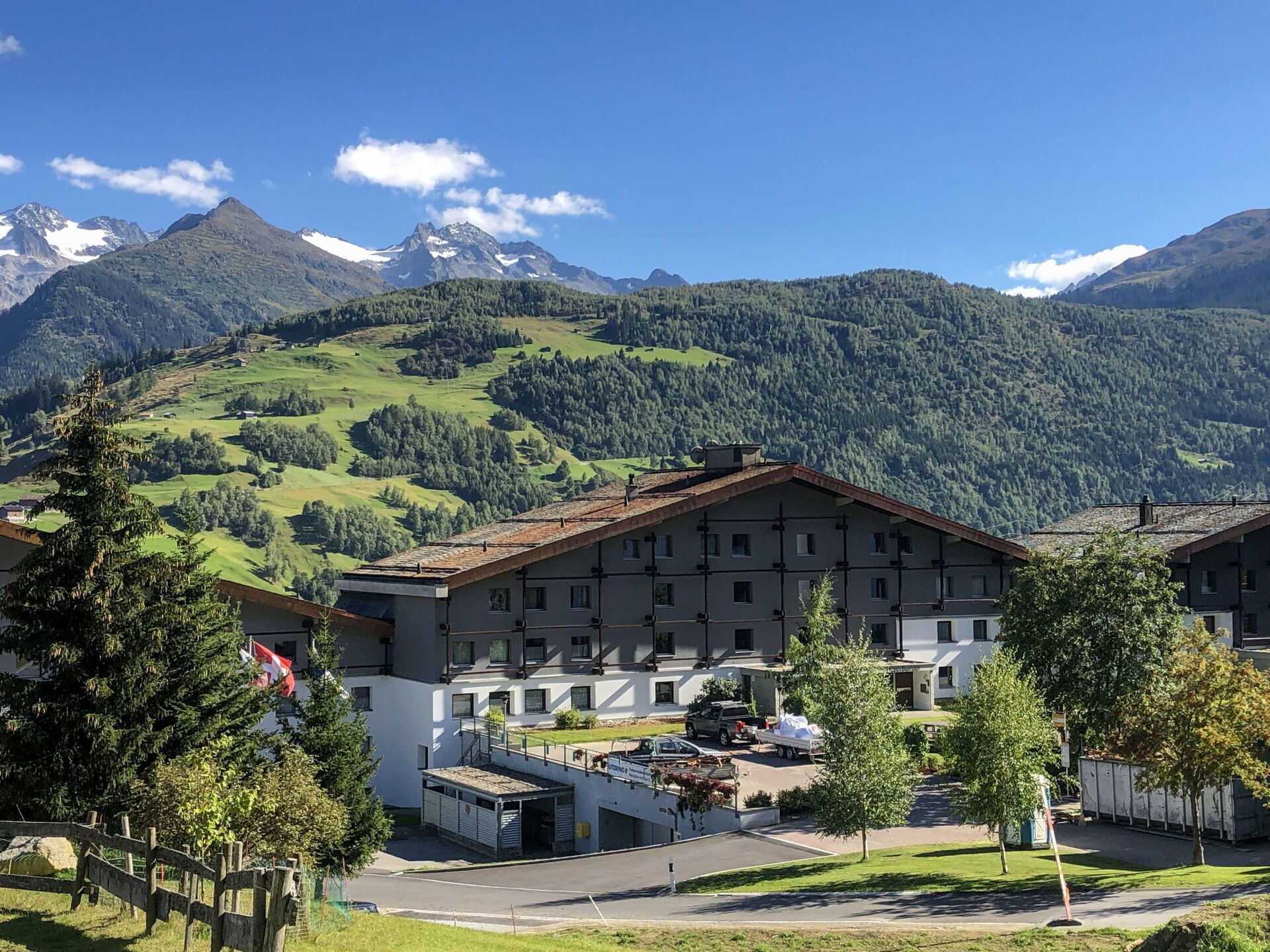 A building with a flat roof and multiple windows, surrounded by greenery and mountains in the background.
