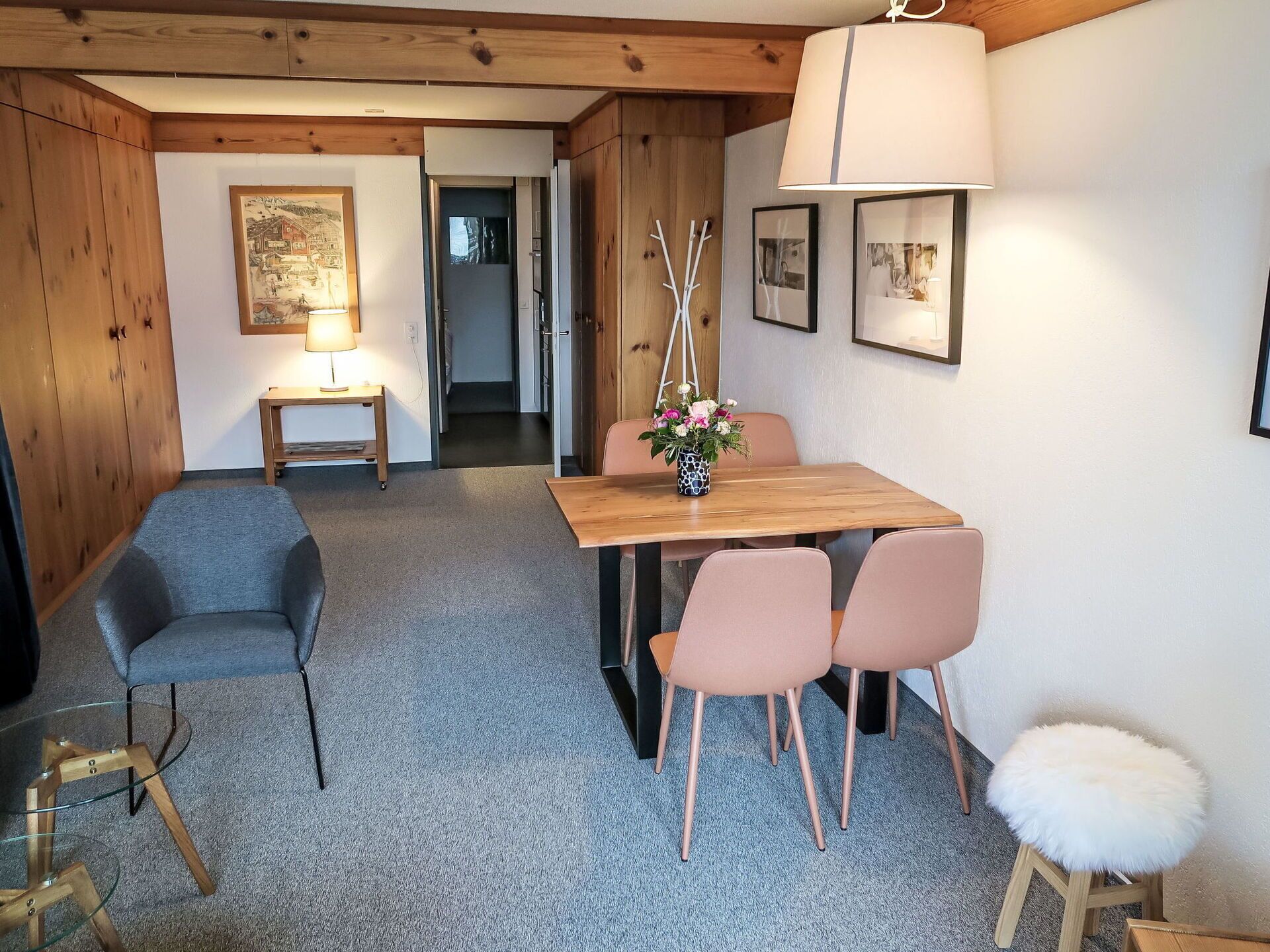 Ein gemütliches Zimmer mit einem Holztisch, zwei Stühlen, einem kleinen Hocker, einer Lampe und einem gerahmten Bild an der Wand.