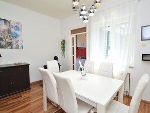 Un espace de salle à manger moderne avec une table et des chaises blanches, un lustre et une vue sur la cuisine, accessible par un passage.