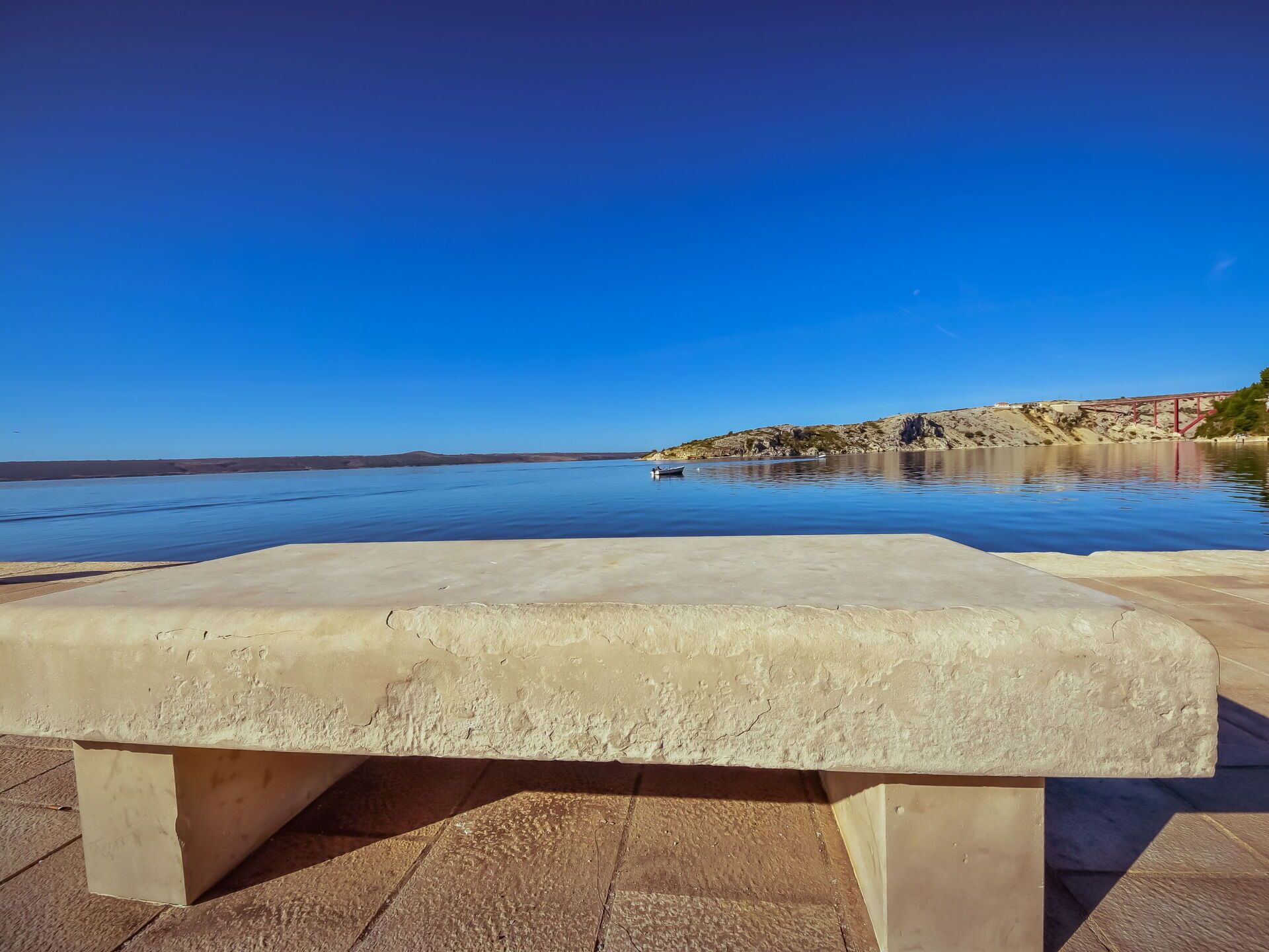 A concrete bench in a public area with a clear view of a calm body of water and a distant rocky shoreline.