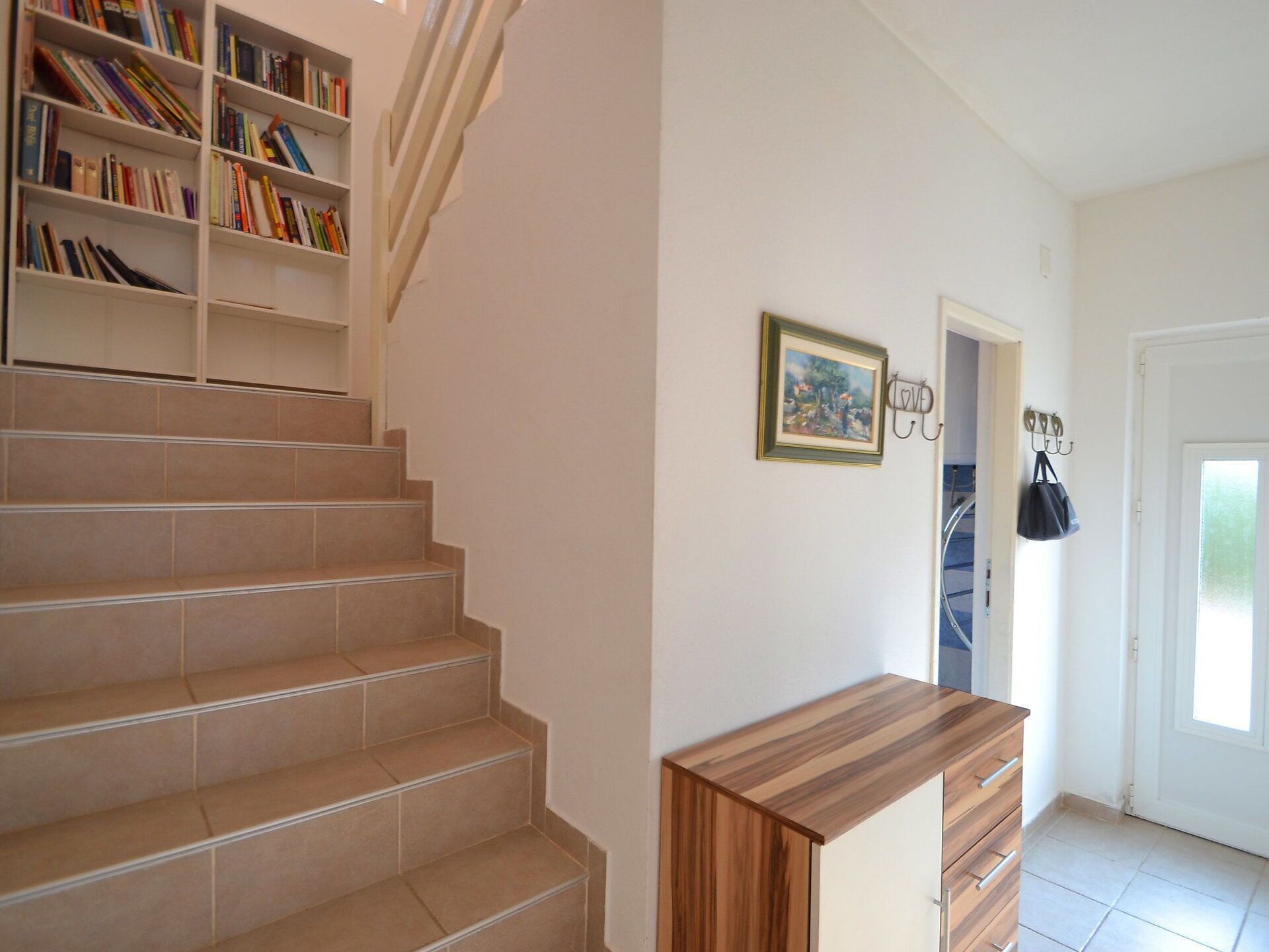 A staircase with beige tiles, a wooden bench, a bookshelf with books, a framed picture, and a coat rack with a bag.