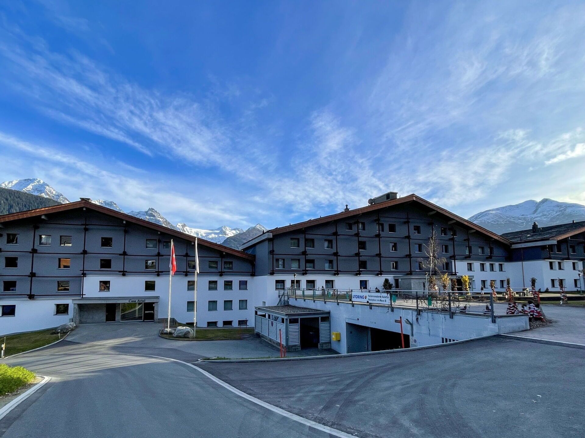 Un hotel moderno con varios pisos, un área de estacionamiento y vistas a montañas nevadas al fondo.