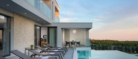 A modern outdoor pool area with lounge chairs and a dining table, surrounded by a stone wall and glass railings.