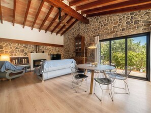 A cozy living room with a stone wall, a fireplace, a dining area with chairs, and a large window offering a view of trees. - Wonderful private villa for 4 people with WIFI, private pool, TV, patio and panoramic view (Inca)