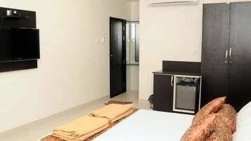 A hotel room with a bed, a flat-screen TV mounted on the wall, a wardrobe, and a small desk with a chair.