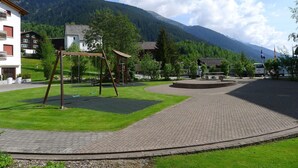 Un terrain de jeux avec des balançoires et une petite maisonnette, entouré d’une zone pavée et de verdure, avec des bâtiments et des montagnes en arrière-plan.