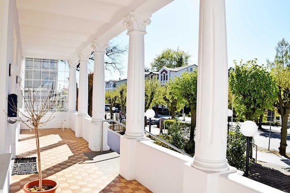 A covered outdoor area with white columns, a tiled floor, and a view of surrounding buildings and trees.
