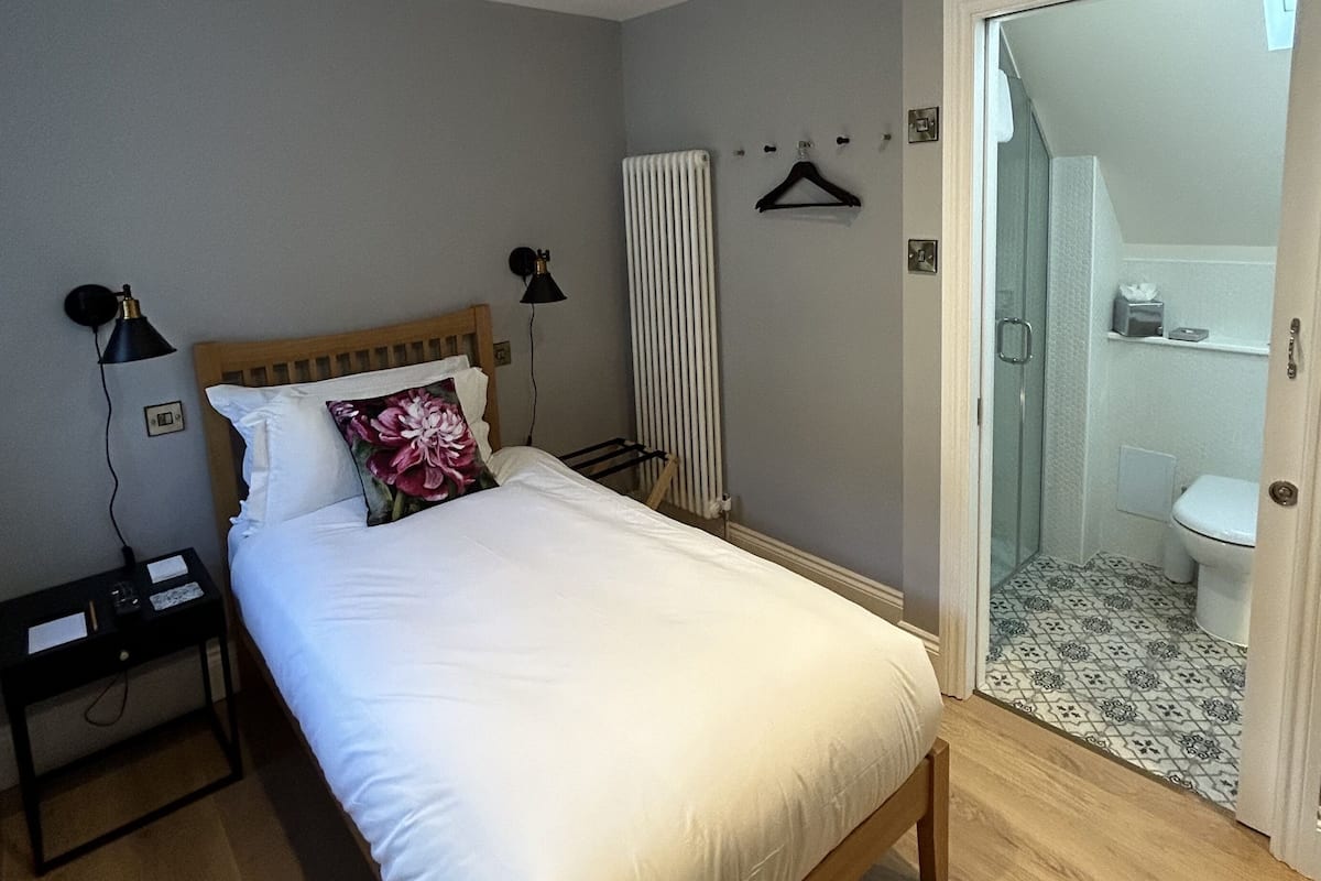 A bedroom with a wooden bed, a nightstand, a lamp, a radiator, and a bathroom visible through an open door.