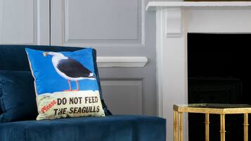 A dark blue sofa with a cushion featuring a seagull and a warning message, positioned in front of a white wall with a gold-framed mirror above a white fireplace.