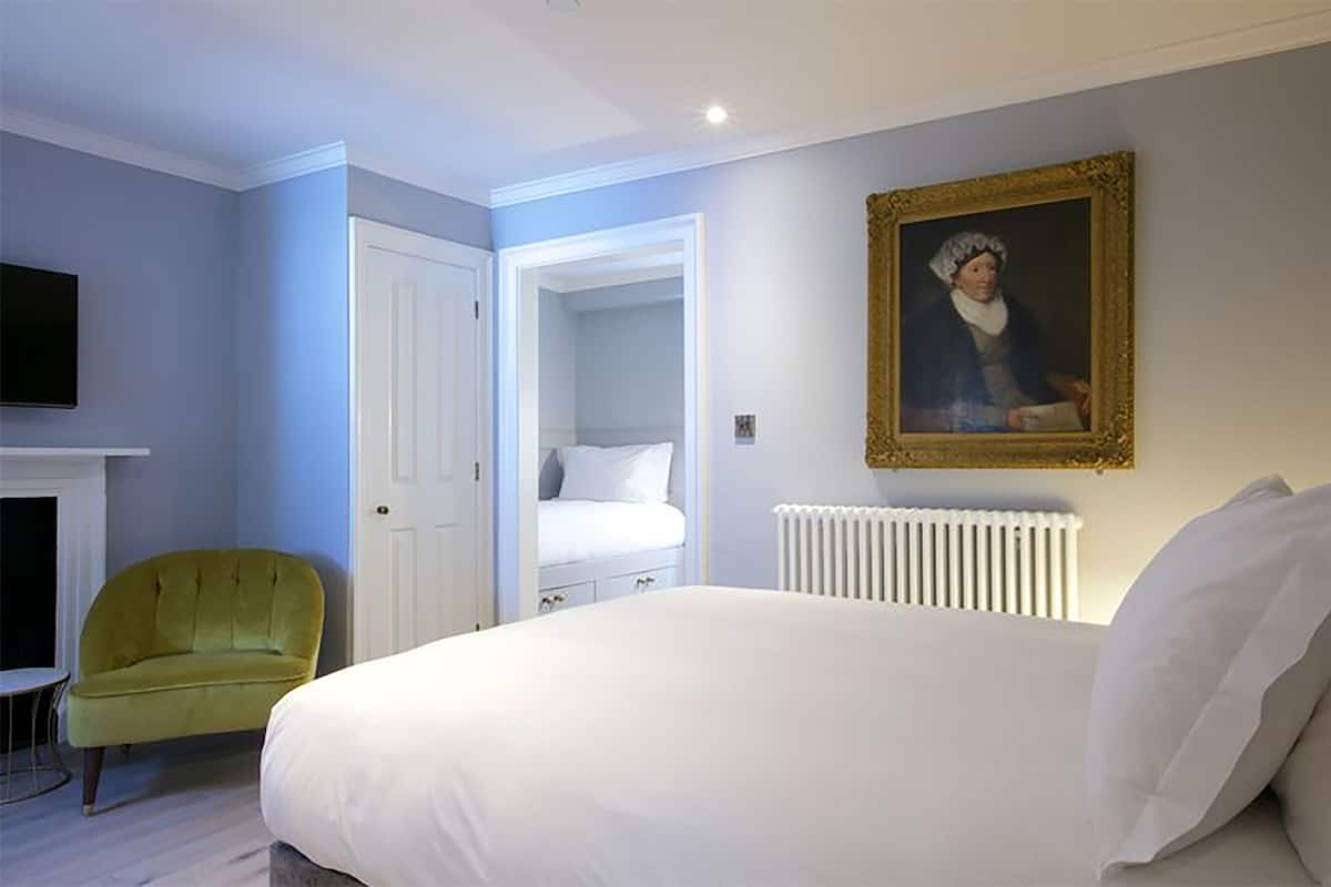 A bedroom with a bed, a green armchair, a white dresser, a painting on the wall, and a fireplace.