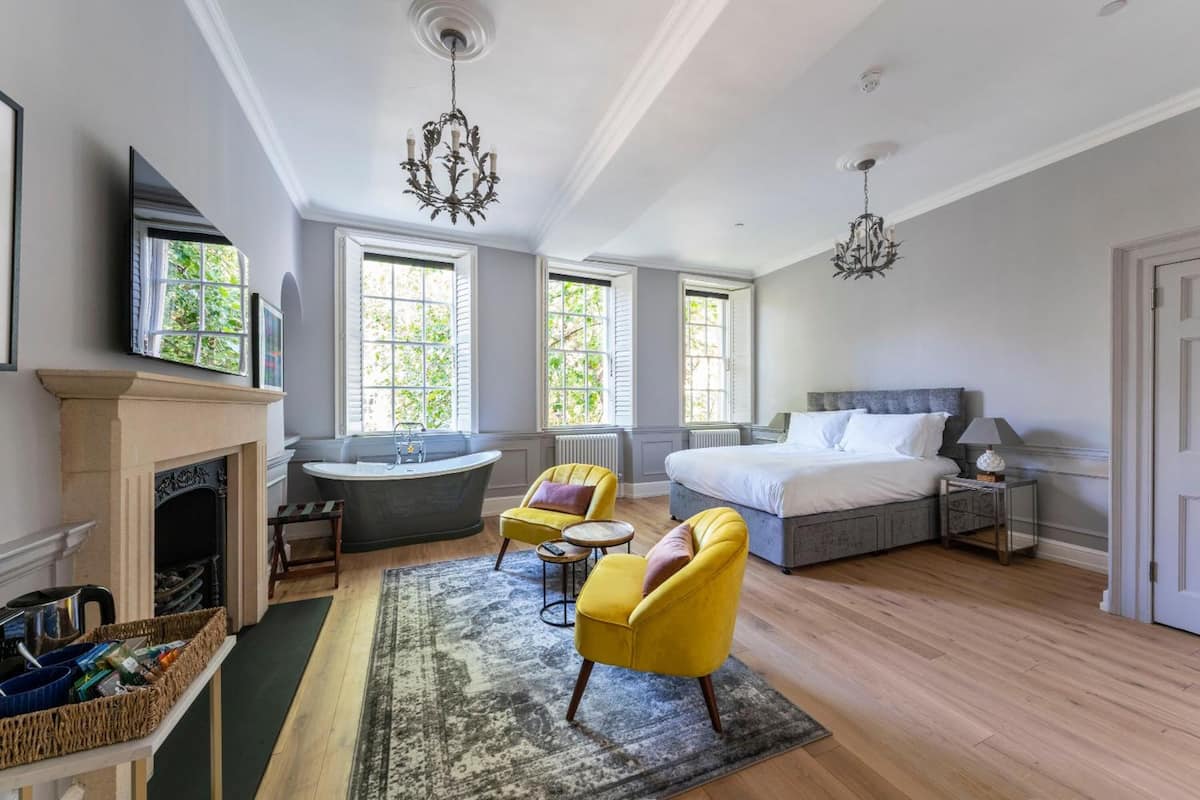 A spacious bedroom with a large bed, a sitting area with yellow chairs, a fireplace, and a view of greenery through the windows.