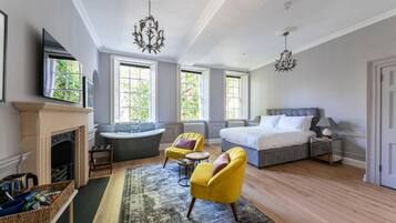 A spacious bedroom with a large bed, a sitting area with yellow chairs, a fireplace, and a view of greenery through the windows.