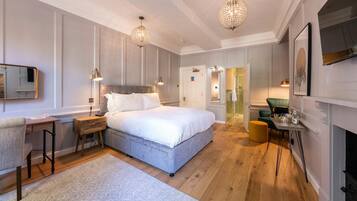 A modern bedroom with a large bed, bedside tables, a desk, and a chair. There are two pendant lights above the bed and a fireplace on the right.