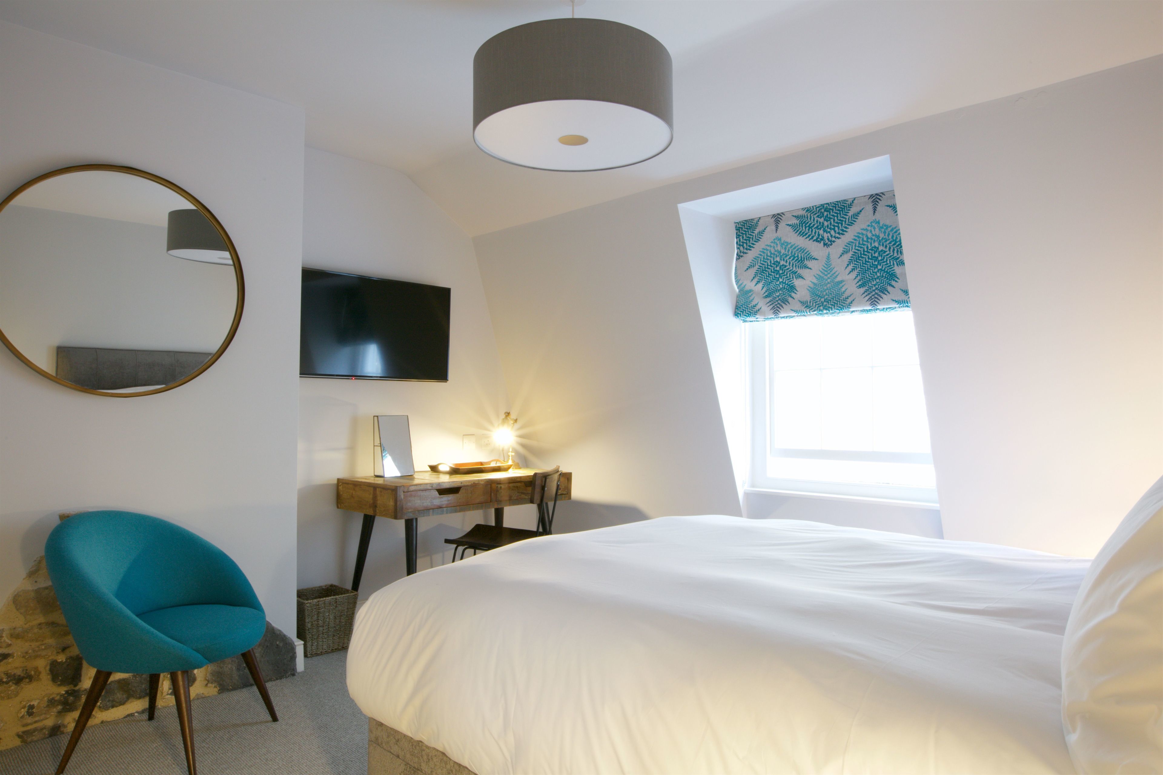 Une chambre à coucher avec un lit, un bureau avec une lampe, une chaise, un téléviseur et une fenêtre avec des rideaux à motifs.