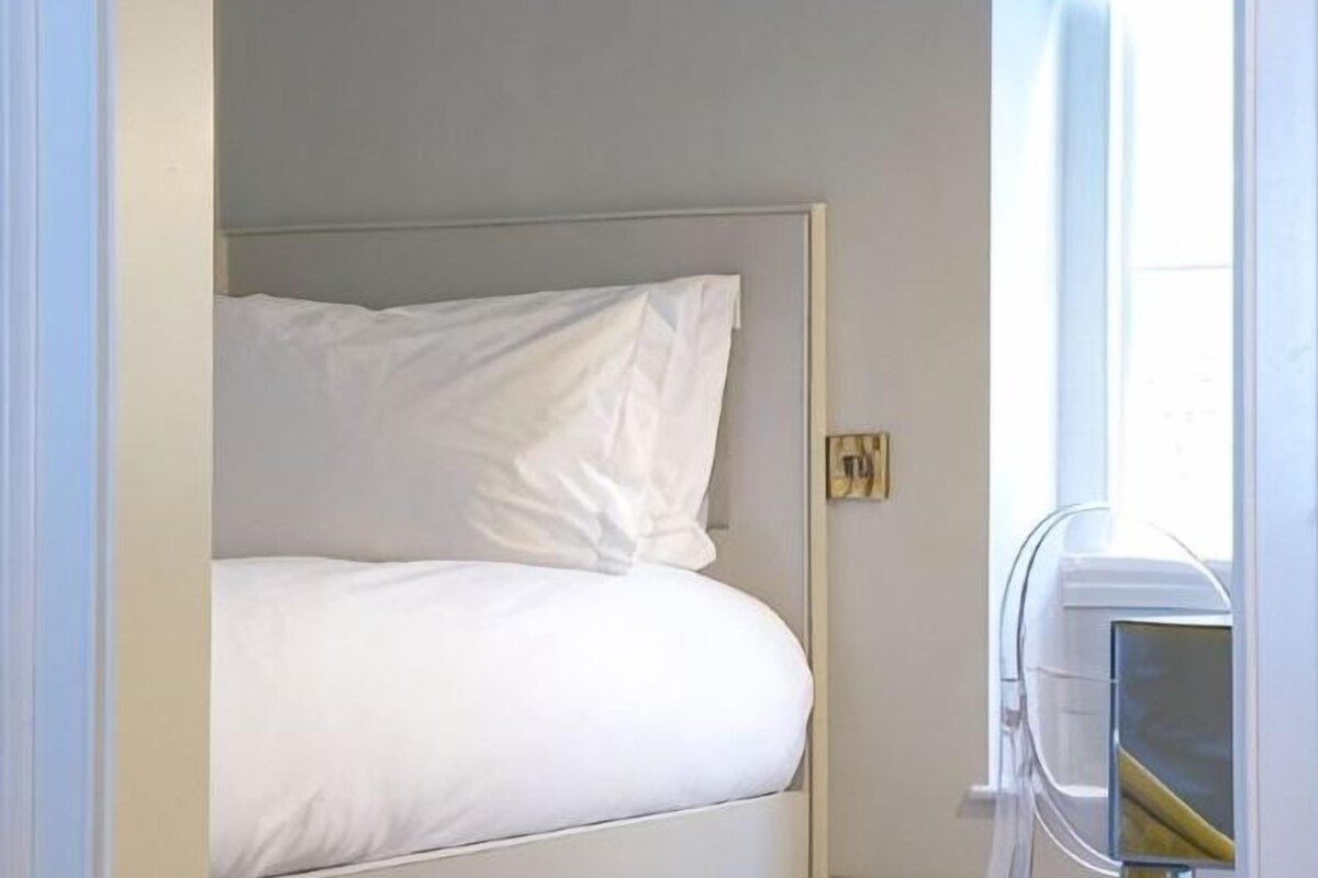 A single bed with white bedding in a room with a plain wall and a window.
