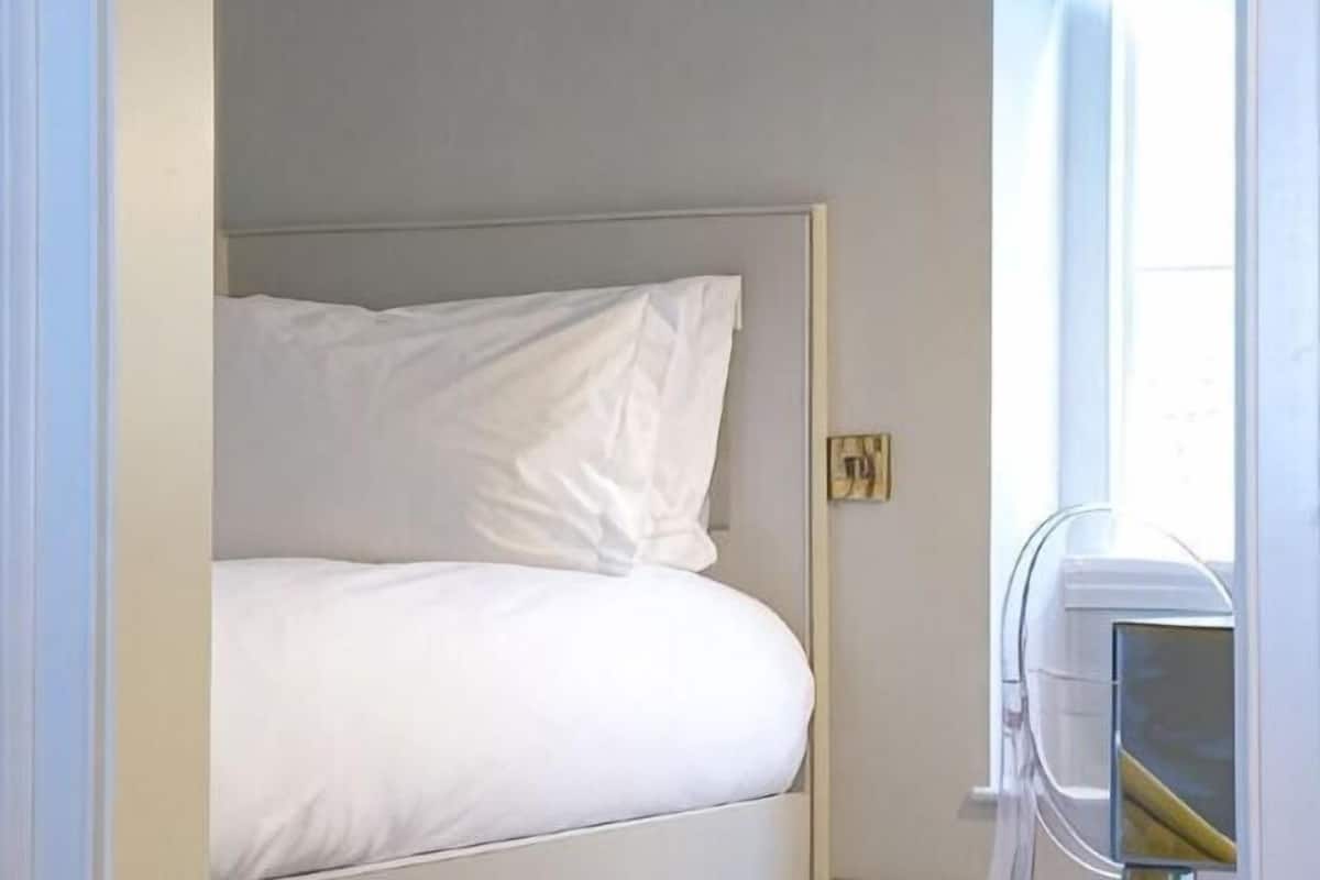 A single bed with white bedding in a room with a plain wall and a window.