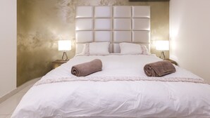 A neatly made bed with a white duvet and two rolled towels, a padded headboard, and two bedside lamps on wooden nightstands.