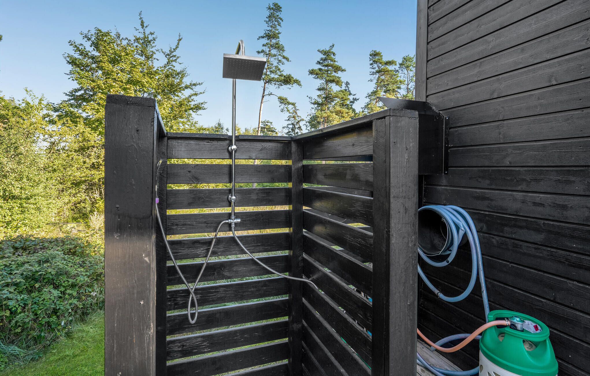 Une cabine de douche extérieure en bois, équipée d’un tuyau d’arrosage et d’une pompe à eau.