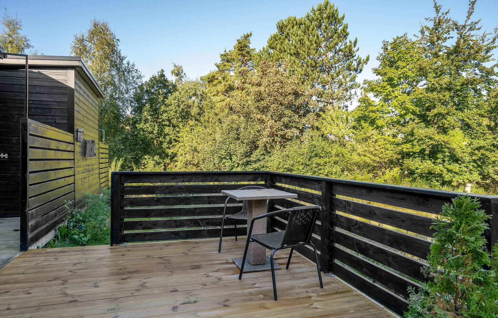 A wooden deck with a table and chairs, surrounded by a black railing, overlooking a forested area.