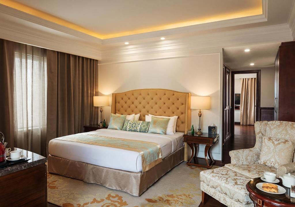 Habitación de hotel con una cama grande, mesitas de noche, un sofá y vistas al pasillo.