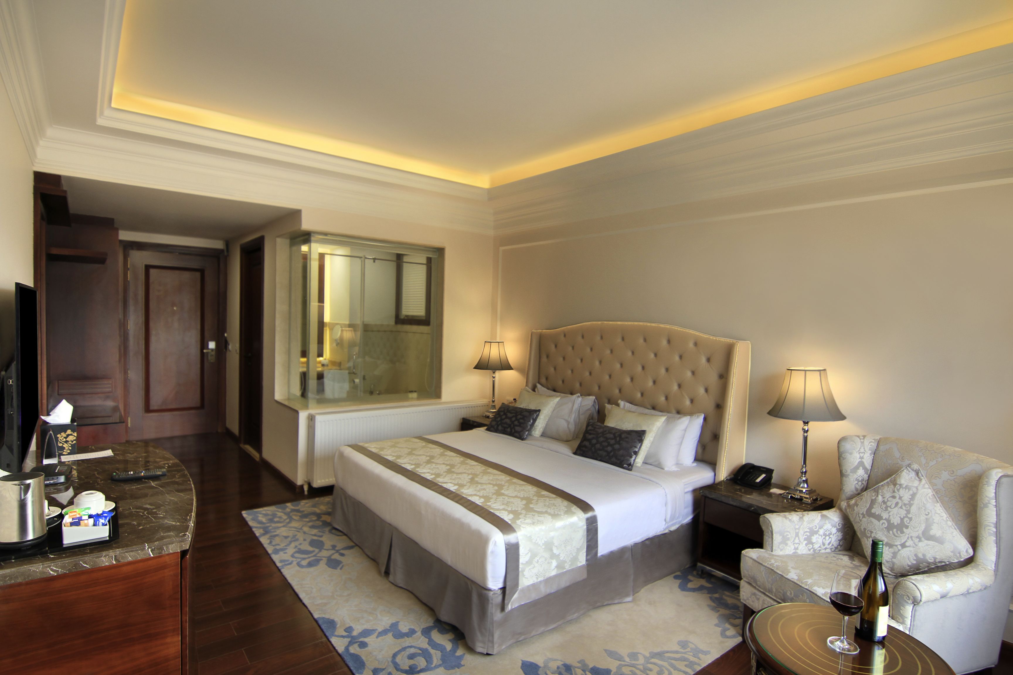 A hotel room with a large bed, bedside tables, a desk with a computer, a chair, and a small table with a bottle and glass.