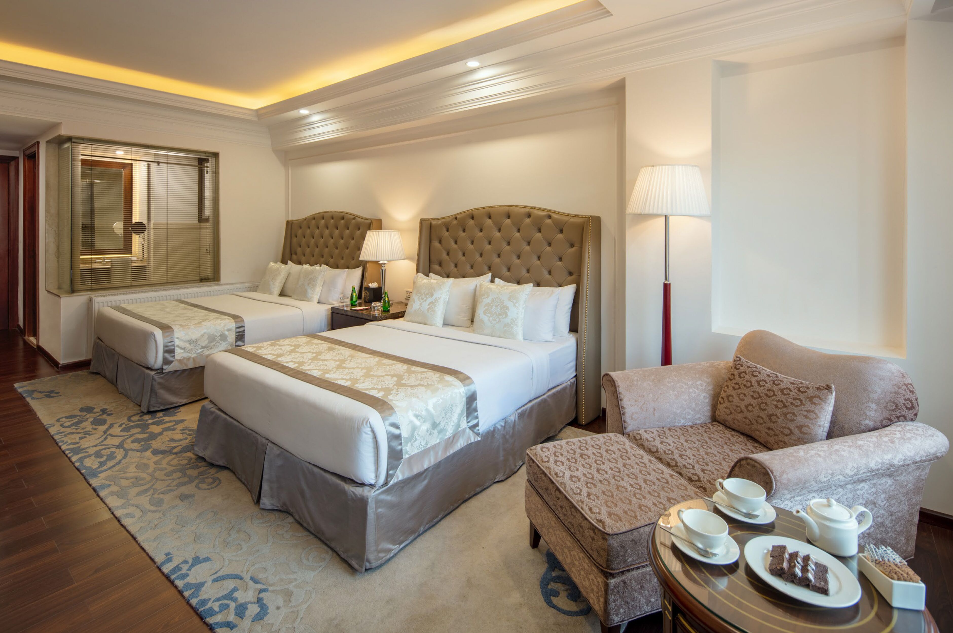 A hotel room with two beds, a sofa, a small table with tea and pastries, and a window with blinds.