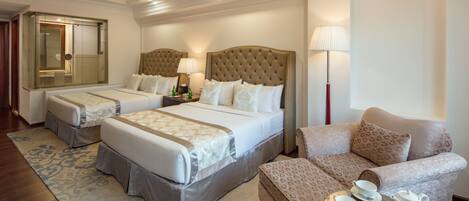 A hotel room with two beds, a sofa, a small table with tea and pastries, and a window with blinds.