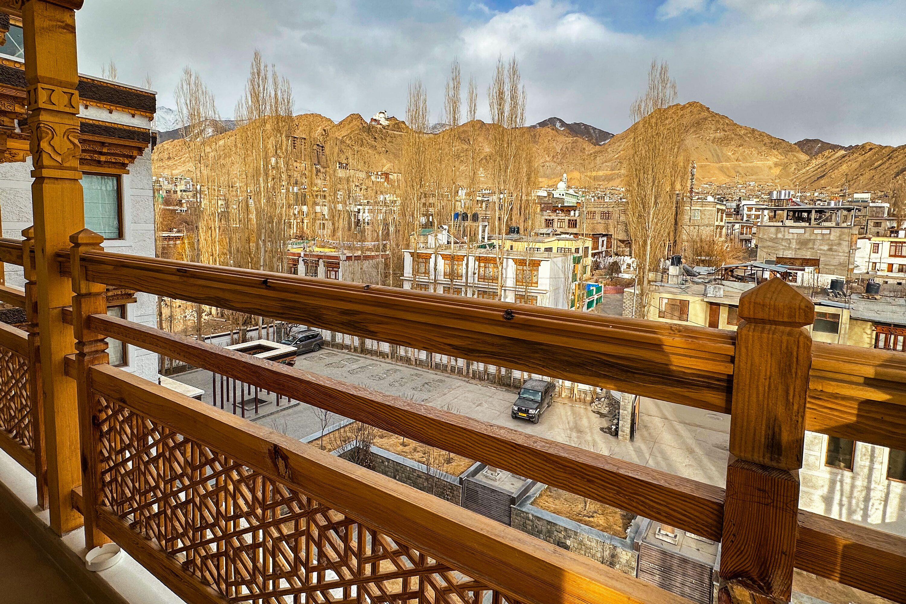A view from a balcony with a wooden railing, overlooking a town with buildings and mountains in the background.