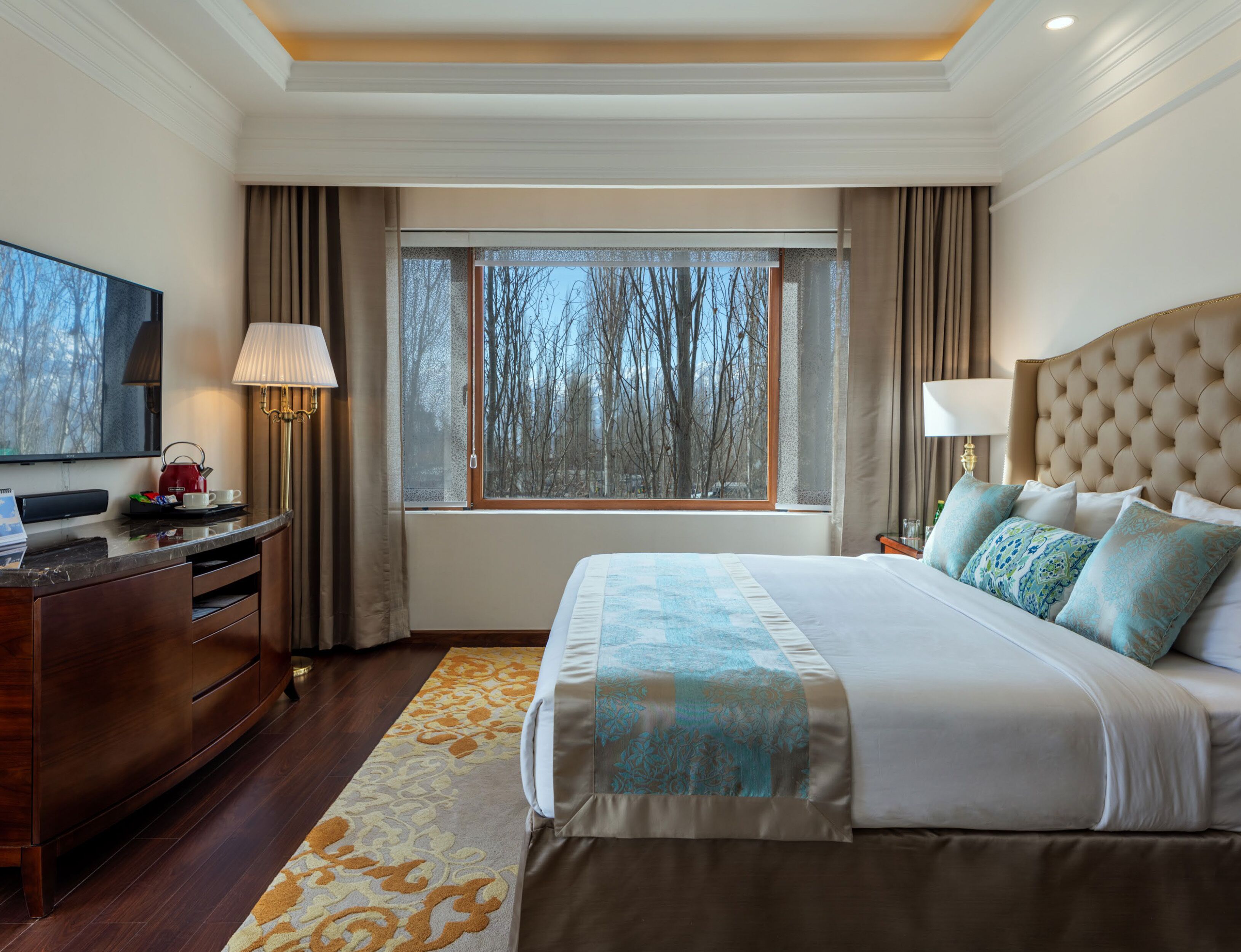 Een hotelkamer met een groot bed, een televisie, een bureau met een lamp en een raam met uitzicht op bomen.