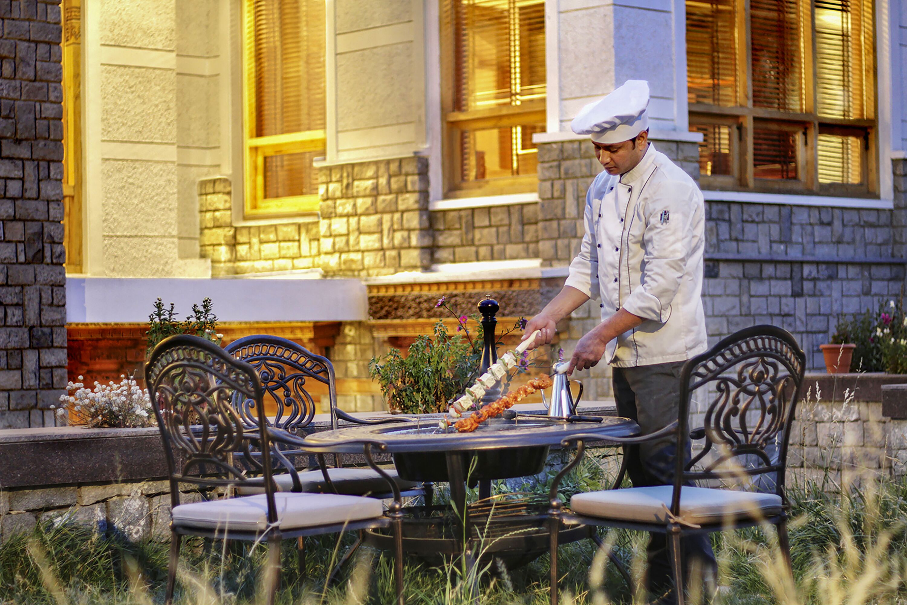 Un chef prépare des plats à l’extérieur, devant une table et des chaises. En arrière-plan, on aperçoit un bâtiment aux murs de pierre et aux fenêtres.