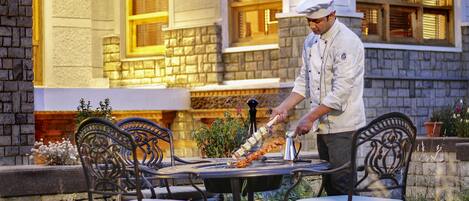 Un chef prépare des plats à l’extérieur, devant une table et des chaises. En arrière-plan, on aperçoit un bâtiment aux murs de pierre et aux fenêtres.
