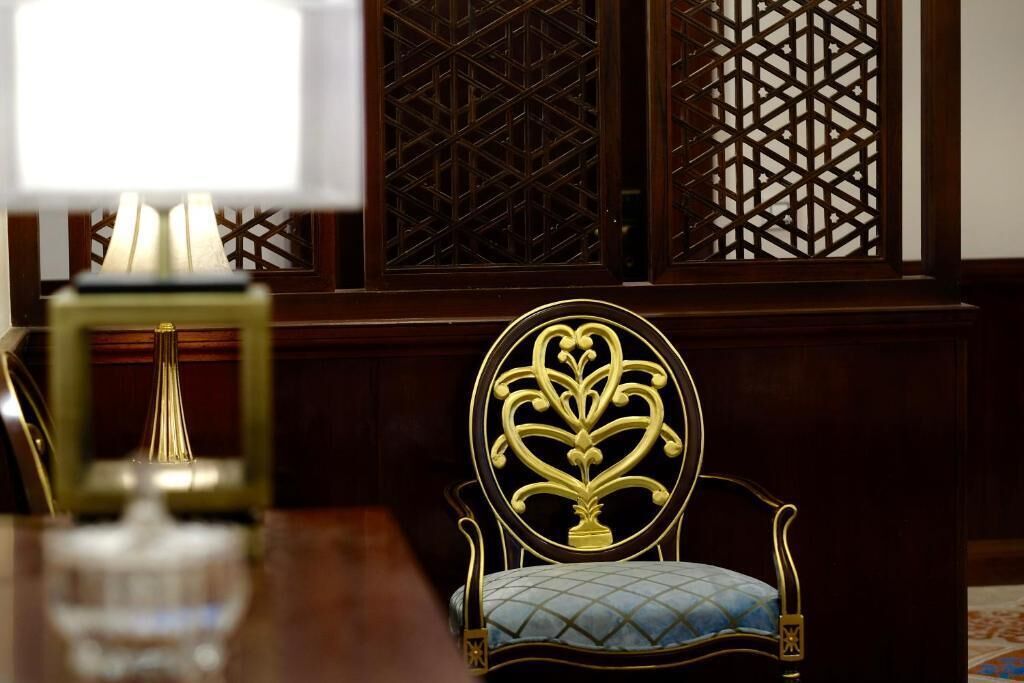 A room with a wooden chair with a gold design, a table with a lamp, and a patterned screen behind the chair.