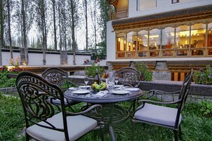 An outdoor dining area with a round table and four chairs, surrounded by greenery and a building with large windows in the background.