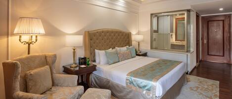 Una habitación de hotel con una cama grande, un área de descanso con una butaca y un reposapiés, una mesita de noche con lámpara, y un espejo amplio.