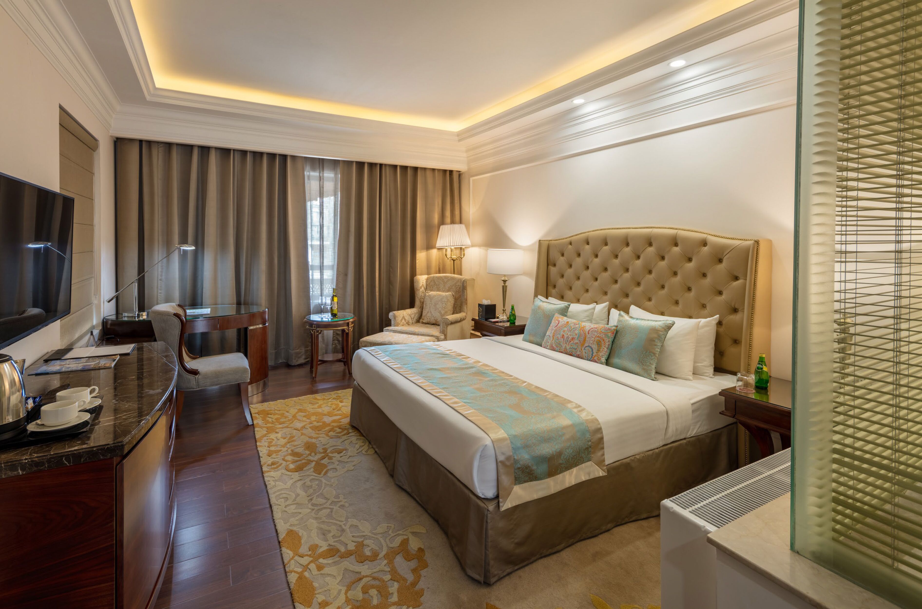 Quarto de hotel com uma cama grande, uma escrivaninha com televisão de tela plana, uma cadeira, um abajur e uma janela com persianas.