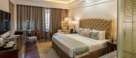 Quarto de hotel com uma cama grande, uma escrivaninha com televisão de tela plana, uma cadeira, um abajur e uma janela com persianas.