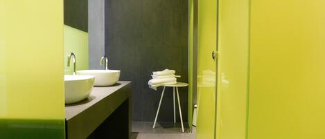 Un baño moderno con dos lavamanos blancos, un taburete pequeño blanco con toallas y una puerta de ducha de cristal.