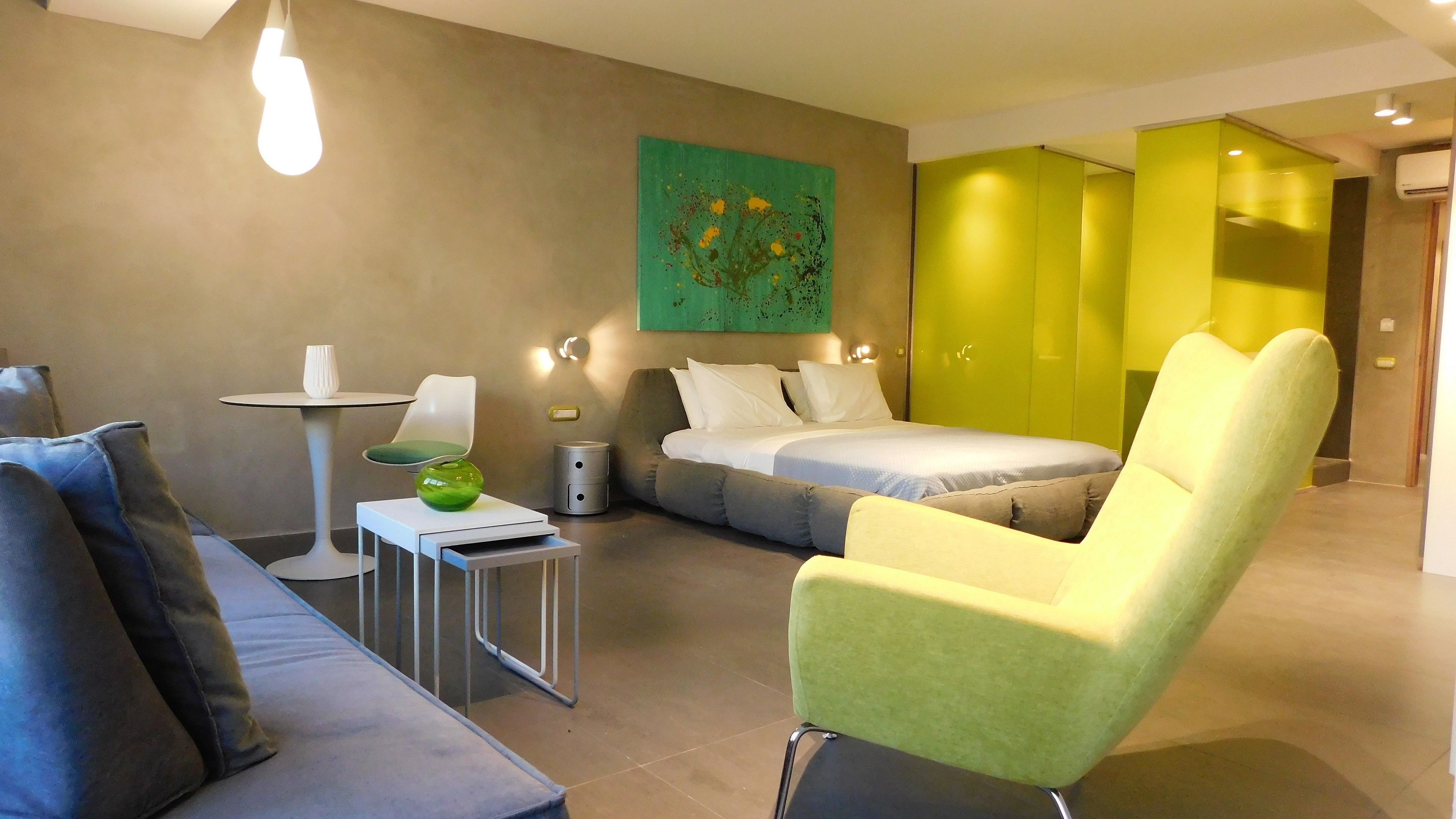 Habitación de hotel moderna con sofá gris, sillón verde, una mesita con un florero, cama con ropa de cama blanca y un cuadro abstracto verde en la pared.