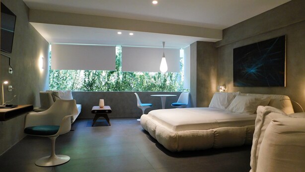 A modern hotel room with a bed, two chairs, a small table, and a view of greenery through a large window.