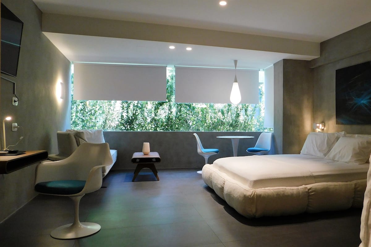 A modern hotel room with a bed, two chairs, a small table, and a view of greenery through a large window.