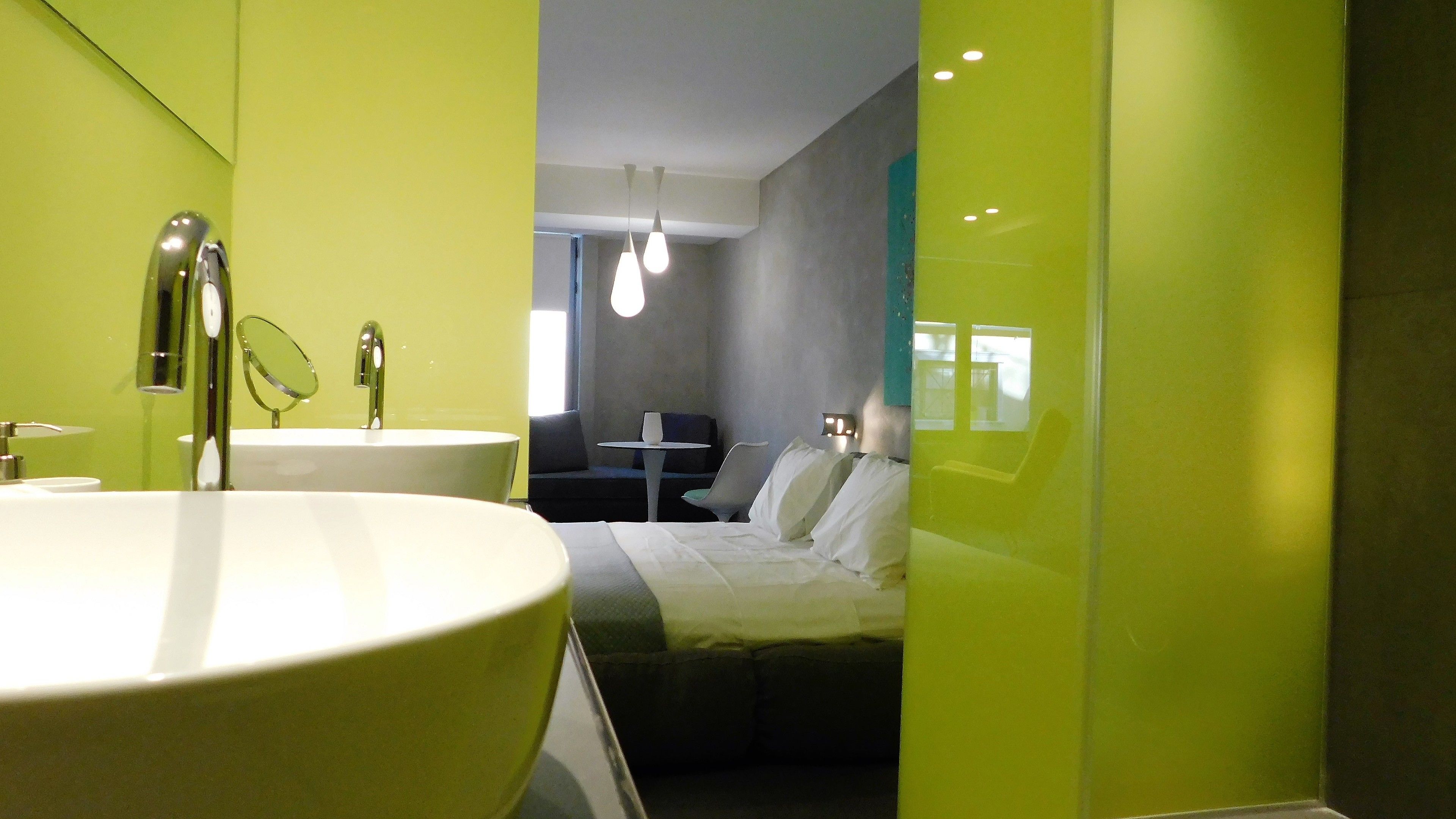 A modern bathroom with a large white sink, a mirror, and a toilet. There is a bed with white bedding and a small table with a chair in the background.