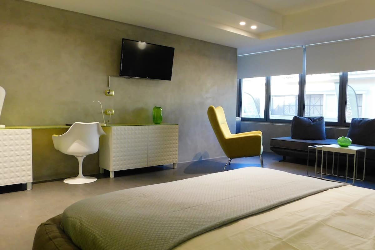 A modern hotel room with a bed, a TV mounted on the wall, a sofa, a chair, and a small table with a green object on it.