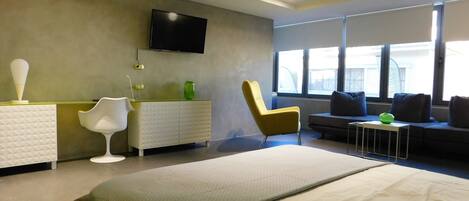Una habitación de hotel moderna con una cama, un televisor montado en la pared, un sofá, una silla y una mesita con un objeto verde encima.