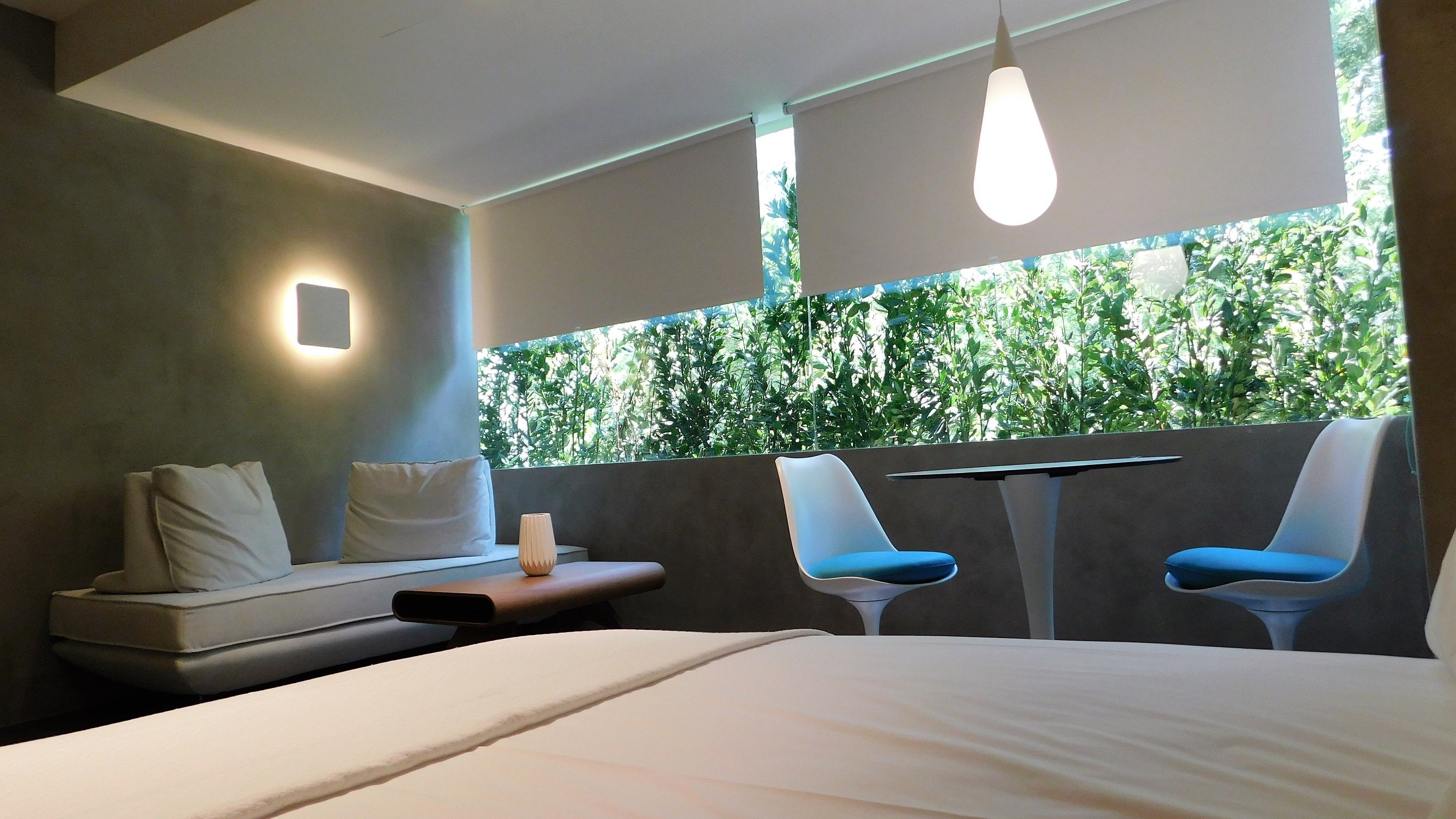 Una habitación de hotel moderna con cama, dos sillas, una mesita y vistas a zonas verdes por una ventana.