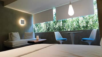 Una habitación de hotel moderna con cama, dos sillas, una mesita y vistas a zonas verdes por una ventana.